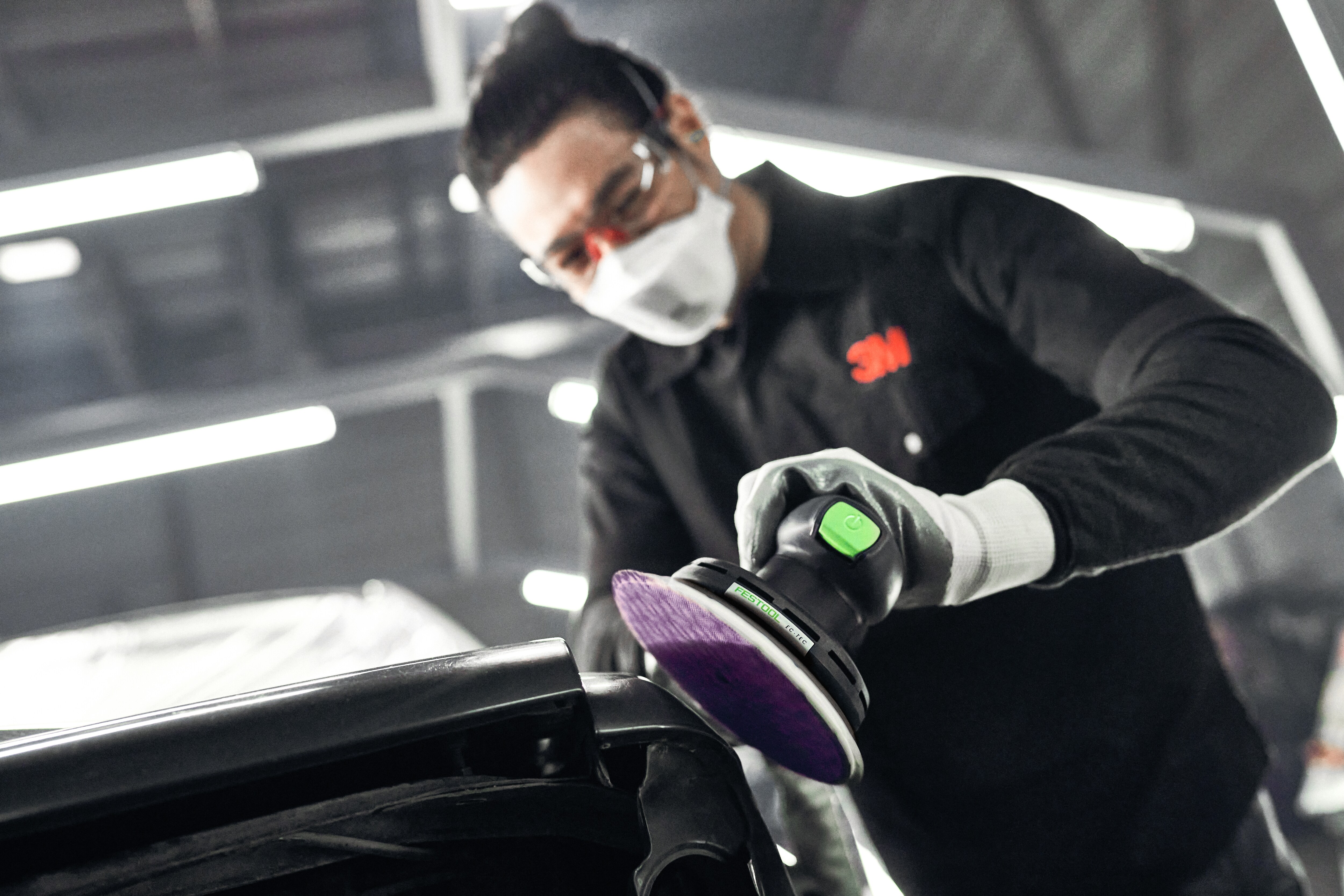 A man wearing safety glasses and a mask using an orbital sander to buff a car bumper.